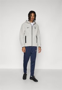 Light grey fleece jacket with a zip closure, black accents, and logo. Paired with navy blue joggers and black athletic shoes.