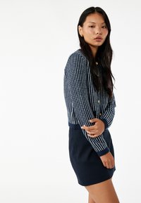 Mörkblå cardigan med vita vertikala ränder, ribbade mudd och krage, i kombination med en enfärgad mörkblå kjol. Materialet verkar vara strukturerat och vävt.