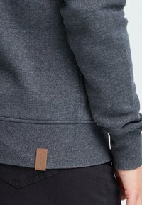 Grijze sweatshirt met een gestructureerde stof, voorzien van een geribde manchetten en zoom. Een klein bruin label is aan de zijkant vlak bij de onderkant gestikt.