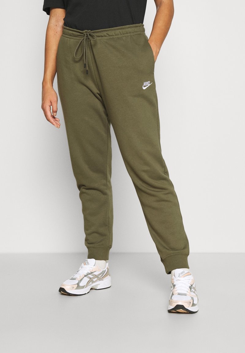 Persoon in olijfgroene Nike-sweatpants met een trekkoord in de taille en witte sneakers, staande tegen een effen lichte achtergrond.