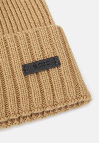 Camel-colored knitted beanie with ribbed texture and a black rectangular label embossed with "BOSS" stitched on the cuff.