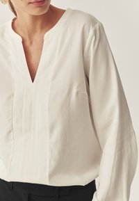 Weiße Bluse mit langen Ärmeln und V-Ausschnitt, die feine Falten und eine einzelne Tasche auf der Vorderseite hat. Glattes Material und lockerer Schnitt.