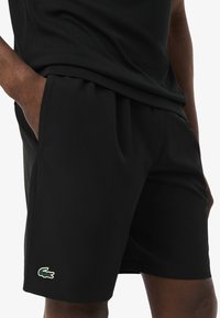 Pantaloni sportivi neri con logo verde Lacoste, indossati da una persona con una mano in tasca, abbinati a una maglietta nera.