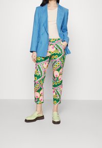 Donna in blazer blu vivo, top beige, pantaloni a fiori vivaci e mocassini verde chiaro in piedi contro uno sfondo semplice.