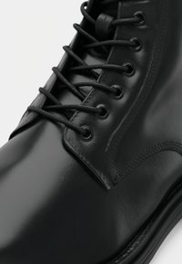 Black leather boot with a glossy finish, featuring round black laces and eyelets. The design includes a reinforced toe and detailed stitching.