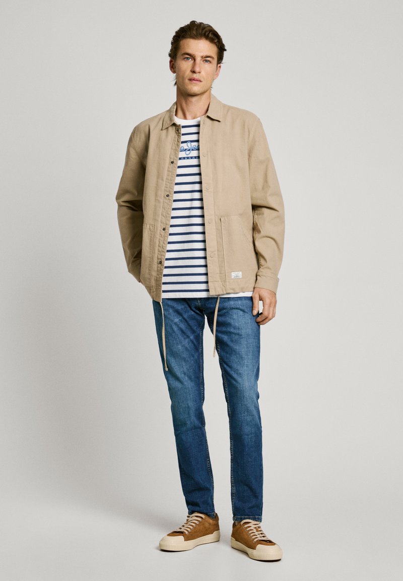 Beige Knopfjacke über einem weiß und marineblau gestreiften T-Shirt, kombiniert mit blauen Jeans und hellbraunen Turnschuhen. Bedeutende Details sind Zugbänder und Taschen.