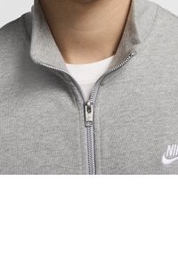 Sudadera gris con cremallera fabricada en un tejido suave, con un cuello alto, cremallera plateada y un logotipo blanco de Nike en el lado izquierdo del pecho.
