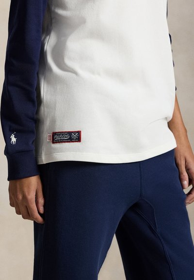 Chemise à manches longues blanche et marine avec logo sur la manche, texture du tissu, et détail de étiquette rouge à l'ourlet ; associée à un pantalon marine.