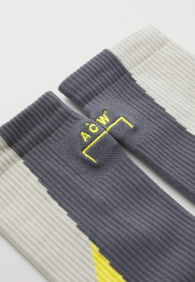 Grå och krämfärgade räfflade strumpor med en gul logopatch. Designen inkluderar färgblockering och en texturerad yta.