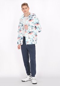 Giacca a motivo floreale bianca con accenti blu e rosa, abbinata a pantaloni blu scuro e sneaker bianche, in piedi su uno sfondo neutro.
