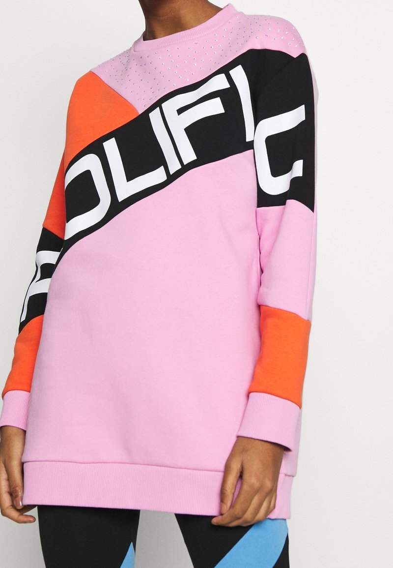 Sudadera rosa con paneles en contraste negro y naranja. Gran texto blanco "POLIFIC" en la parte frontal, con acentos de strass en los hombros.