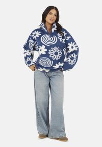 Donna in piedi con le mani in tasca, che indossa un pullover di pile blu e bianco con un motivo floreale e jeans a gamba larga di un blu chiaro, abbinati a scarpe beige.