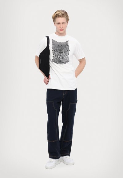 Balta kokvilnas T-kreklā ar melnām viļņainām līnijām, apvienota ar tumši zilām kravas biksēm ar redzamām šuvēm un baltām sporta kedām. Melna plecu soma.