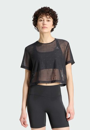 Žena s krátkými kudrnatými vlasy, která má na sobě černý průsvitný crop top přes černou sportovní podprsenku a černé vysoce střižené cyklistické šortky, stojí.