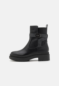 Botas de tobillo de cuero negro con suela gruesa, que presentan una correa de tela decorativa y un acento de logo en el lateral. Detalle de panel texturizado.