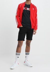 Giacca da track rossa con zip e strisce bianche, t-shirt nera con il testo "BORED", pantaloni corti neri, calzini bianchi con strisce nere e sneakers bianche.