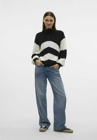Sweter w kolorze czarnym i białym w paski, z golfem i zamkiem błyskawicznym, w zestawie z niebieskimi spodniami typu wide-leg i szarymi trampkami z czarnymi sznurówkami.