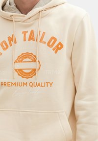 Beige Kapuzenpullover mit vorderer Eingrifftasche, Kordelzug-Kapuze und orangener Grafikschrift. Weicher Stoff und lässiges Design. Mit "Premium Quality"-Branding.
