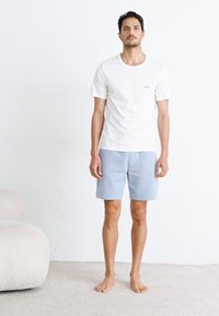 Witte t-shirt met korte mouwen en een klein logo, gecombineerd met lichtblauwe gestreepte shorts. Model staat blootsvoets op een licht tapijt.
