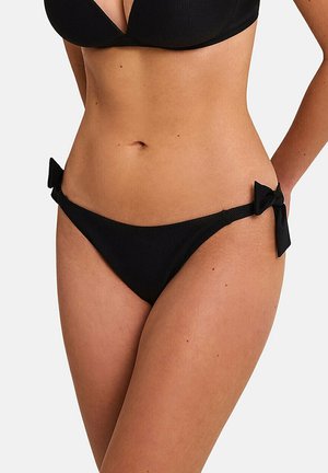 Vrouw met een zwarte bikinibroek met zijstrikjes en een bijpassende zwarte bikinitop, staand tegen een effen lichte achtergrond.