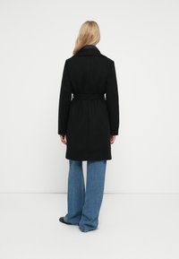 Cappotto nero con vita cinturata, colletto a punta e maniche lunghe; abbinato a jeans blu a gamba larga. Tessuto liscio e design su misura.