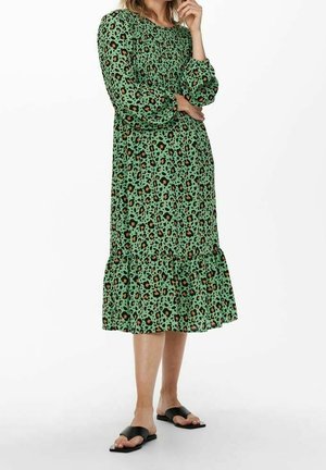 Frau trägt ein grünes Midikleid mit Leopardenmuster, langen Puffärmeln und schwarzen flachen Sandalen, steht mit einer Hand nahe am Gesicht.