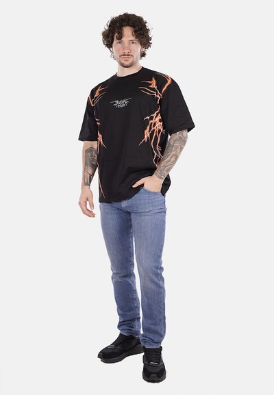 T-shirt nera a maniche corte con grafiche di fulmini arancioni. Tessuto in cotone, vestibilità rilassata. Indossata con jeans azzurri chiari e sneakers nere.