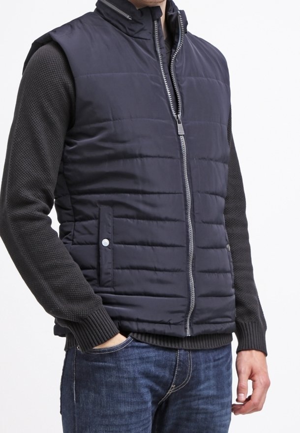 Granatowa puchowa kamizelka z pikowanym wzorem, zapinana na zamek, z bocznymi kieszeniami, zestawiona z teksturowaną czarną koszulą z długim rękawem i niebieskimi dżinsami.