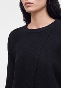 Femme aux longs cheveux foncés portant un pull noir texturé à col rond sur un fond clair uni.