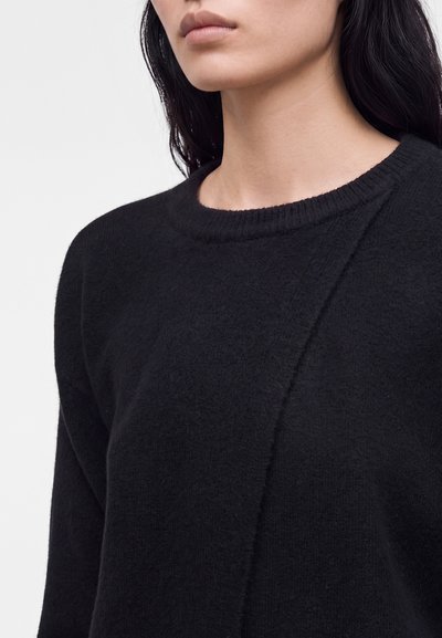 Femme aux longs cheveux foncés portant un pull noir texturé à col rond sur un fond clair uni.