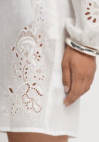 Vit linneklänning med detaljerad broderad snitt längs sidan. Slät textur, långa ärmar och ett silverarmband på handleden.