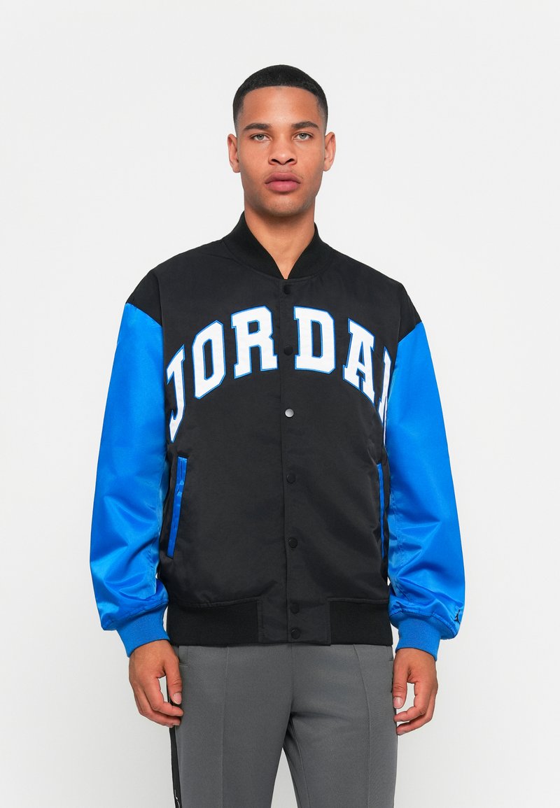 Black and blue varsity jacket featuring bold white "JORDAN" lettering, a snap-button front, and ribbed cuffs. Smooth fabric with side pockets.