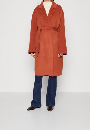 Persona che indossa un cappotto color ruggine con cintura, un maglione bianco, jeans blu e scarpe nere a punta su uno sfondo chiaro e semplice.