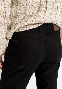 Pantaloni in denim nero con una texture liscia e tasche posteriori cucite. Indossati con un maglione spesso a trama a trecce color crema.