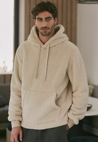 Beige fleece-huppari, jossa on teksturoitu pinta, naruvetoketjuhuppu ja etutasku. Ominaisuuksiin kuuluu rento malli ja pitkät hihat.
