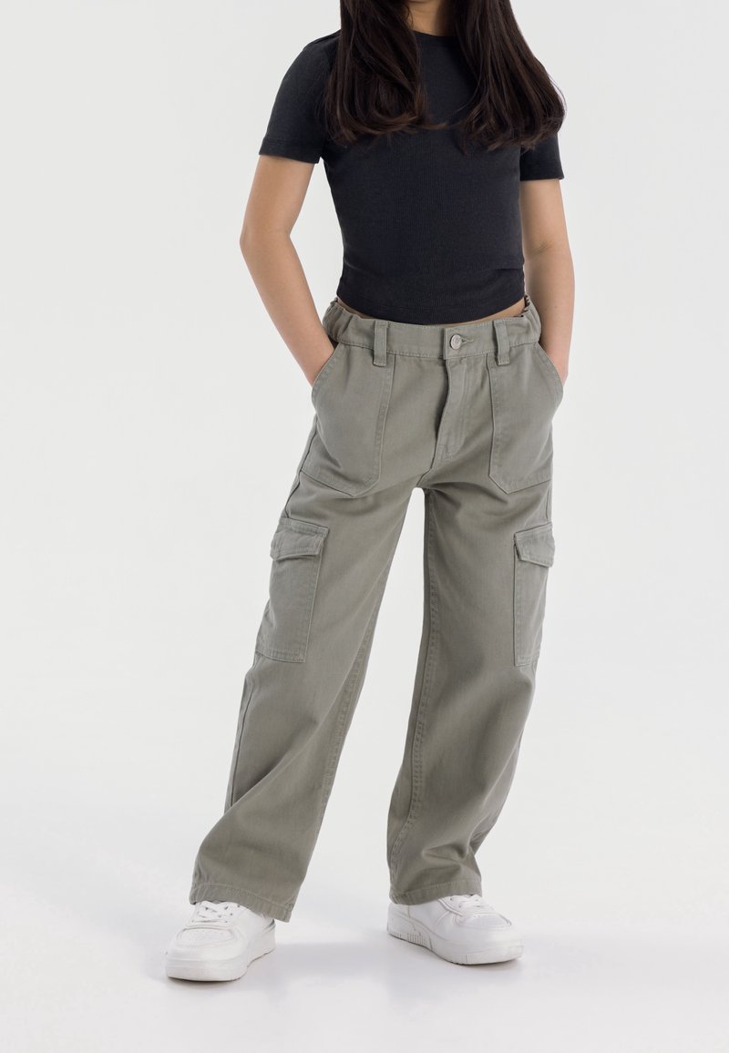 Personne portant un haut noir à manches courtes, un pantalon cargo gris clair ample avec de grandes poches et des baskets blanches, debout les mains dans les poches.