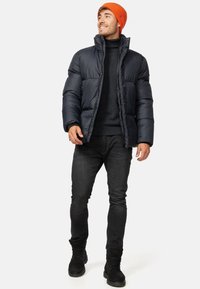 Marineblå pufferjakke med høj krave, parret med en sort turtleneck, mørke jeans og sorte støvler. Lys orange strikbeanie tilføjer kontrast.