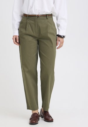 Femme portant un pantalon fuselé taille haute vert olive avec une ceinture marron, une chemise blanche, un bracelet noir et des mocassins marron, debout sur un fond blanc.