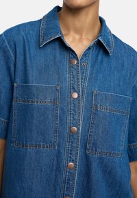 Niebieska koszula dżinsowa z klasycznym kołnierzykiem, krótkim rękawem, dwoma przednimi kieszeniami i brązowymi guzikami przy zapięciu.