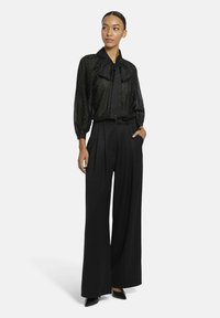Zwarte, transparante blouse met een strik om de hals en een metalen stippen patroon; gecombineerd met hooggewaiste, wijde zwarte broeken en puntige schoenen.