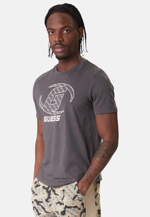 Uomo con capelli intrecciati che indossa una T-shirt grigia con il logo Guess e pantaloncini beige a motivo, in piedi con le mani nelle tasche contro uno sfondo bianco.