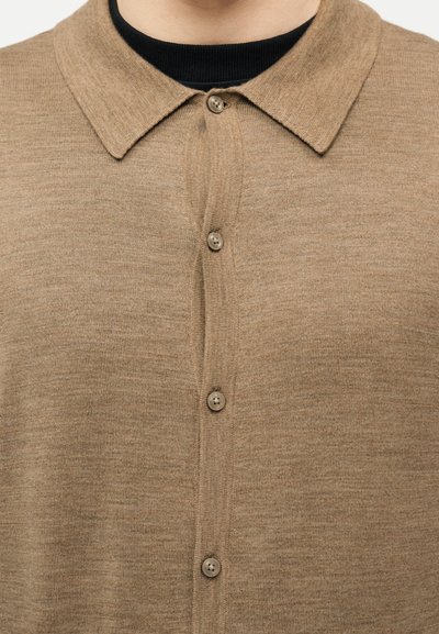 Μπεζ πουκάμισο με κουμπιά και μυτερό γιακά, φορεμένο πάνω από μαύρο μπλουζάκι με στρογγυλή λαιμόκοψη, εμφανιζόμενο από το μέσο του στήθους έως την περιοχή του λαιμού.