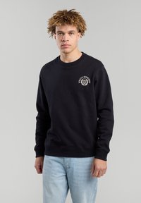 Black sweatshirt made from cotton, featuring long sleeves, a crewneck, and a small white graphic on the left chest. Worn with light blue jeans.