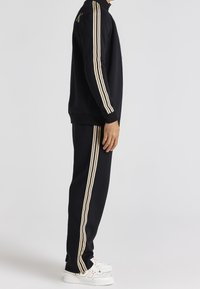 Chándal negro con rayas beige en las mangas y pantalones, con un cuello alto y puños acanalados. Completado con zapatillas blancas.