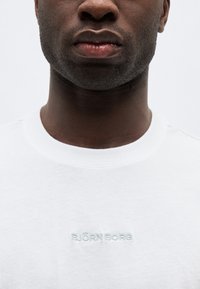 Κοντινή λήψη του κάτω μέρους του προσώπου και του λαιμού ενός άνδρα που φορά λευκό πουκάμισο με το κέντημα "BJÖRN BORG" στο στήθος.