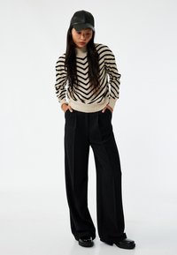 Beiger Pullover mit schwarzen Zickzackstreifen, hoher Kragen; schwarze weit geschnittene Hose mit Taschen; glänzende schwarze Schuhe; schwarze Lederkappe.