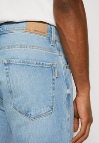 Los jeans de mezclilla de color azul claro presentan un parche de cuero en la cintura etiquetado como "ANTONY MORATO", con un diseño estándar de cinco bolsillos y costuras visibles.