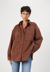 Giacca camicia in corduroy marrone con neckline a colletto, due tasche sul petto e vestibilità ampia. Presenta una texture a costine e chiusura a bottoni.