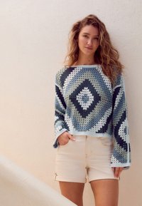 Sweter szydełkowy w kolorze niebieskim i białym z wzorem diamentowym, o luźnym kroju i szerokich rękawach. Połączony z jasnobeżowymi szortami.