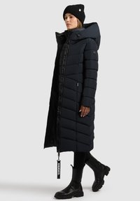 Sacou puffer lung, negru, cu fermoar frontal, model matlasat în formă de zigzag și glugă supradimensionată, purtat peste colanți negri și botine.